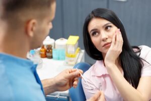 Woman in dentist’s chair holding mouth in pain with attending dentist nearby.